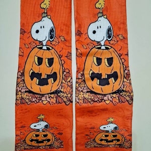 以下が含まれることがあります： Snoopyとウッドストックが描かれたオレンジ色のハロウィン柄の靴下2足セット。 スヌーピーはカボチャの衣装を着ており、ウッドストックが頭に乗っています。 靴下にはオレンジと茶色の葉の模様があります。