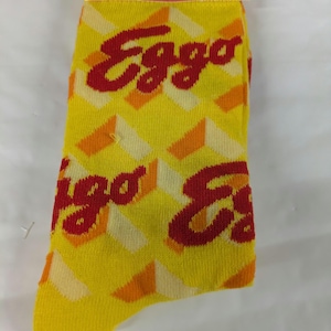 May include: Yellow crew socks with the Eggo logo in red. The socks have a repeating pattern of yellow, orange, and white geometric shapes. The packaging reads "Eggo Crew Socks" and "Fits Men's Shoe Size 6-12."