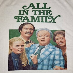 May include: A white t-shirt with a green print of the words "ALL in the FAMILY". The print also includes a photo of the four main characters from the TV show.