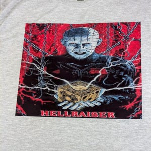 May include: A light gray t-shirt featuring a graphic from the movie Hellraiser. The graphic shows a character holding a puzzle box, with the word "HELLRAISER" in red at the bottom. The background is red with chains and lightning.