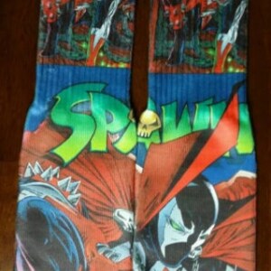 May include: A pair of blue and green socks with a graphic print of the comic book character Spawn. The socks feature the words "Spawn" in a green and white font.