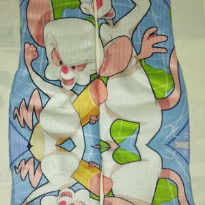Può includere: Un paio di calzini con i personaggi dei cartoni animati Pinky e il Cervello. I calzini hanno uno sfondo blu con un disegno della mappa del mondo e illustrazioni dei due topi. Il testo "Pinky and The Brain" è visibile.