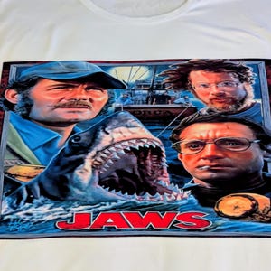 Może przedstawiać: Biała koszulka z motywem plakatu filmowego. Projekt zawiera słowo "JAWS" oraz wizerunki postaci z filmu i rekina. Grafika plakatu jest oprawiona w ciemnoczerwoną ramkę.
