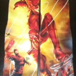 May include: A pair of black socks with a colorful graphic print of Daredevil, a superhero known for his red suit and billy clubs. The socks feature multiple images of Daredevil in different poses, including one with a red and yellow background.
