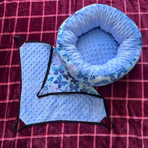 May include: A round, light blue pet bed with a textured dimpled surface and a butterfly print on the exterior, paired with a matching square hammock featuring black trim and straps, both made from soft, plush fabric with a consistent blue color.
