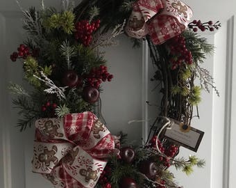 May include: A Christmas wreath with a red and white plaid ribbon with gingerbread men and candy canes. The wreath is decorated with red berries, green pine branches, and red apples.