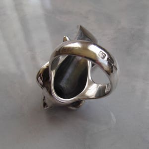 May include: A silver ring with a detailed design of a stylized animal head. The ring is open on the side and has a black stone in the center. The ring is marked with the number "925" on the inside of the band.
