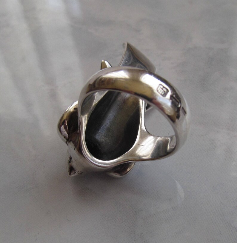 May include: A silver ring with a detailed design of a stylized animal head. The ring is open on the side and has a black stone in the center. The ring is marked with the number "925" on the inside of the band.