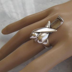 May include: A silver ring with a unique abstract design, resembling a stylized bird or flower. The ring is worn on a person's finger.
