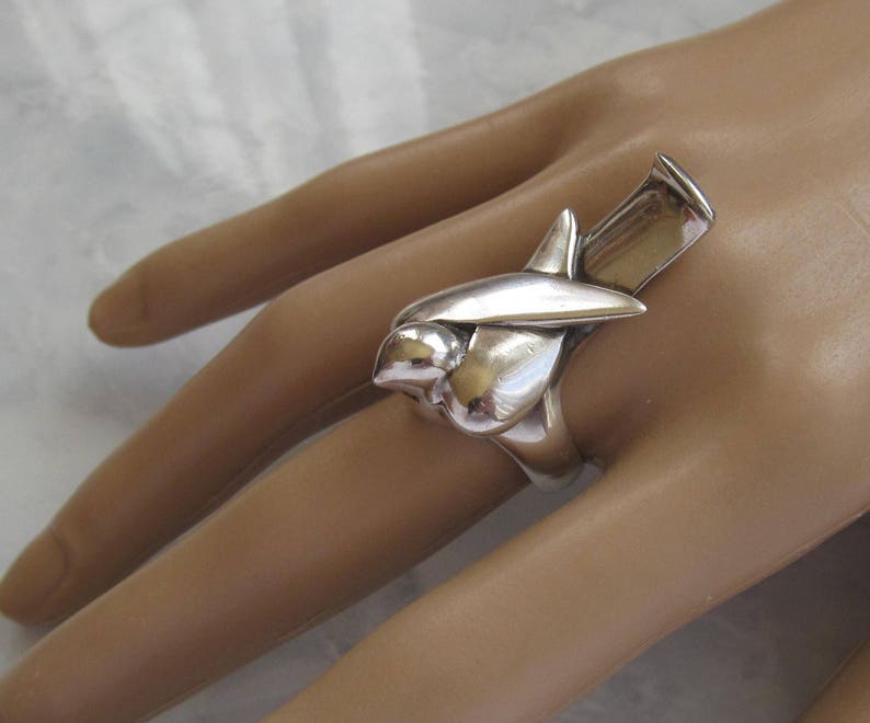 May include: A silver ring with a unique abstract design, resembling a stylized bird or flower. The ring is worn on a person's finger.