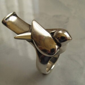 May include: A silver ring featuring a stylized bird design. The bird is perched on a branch, with its wings spread out. The ring is made of a smooth, polished metal.