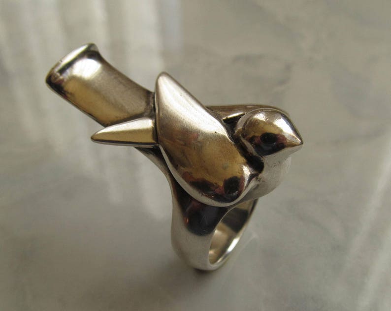 May include: A silver ring featuring a stylized bird design. The bird is perched on a branch, with its wings spread out. The ring is made of a smooth, polished metal.
