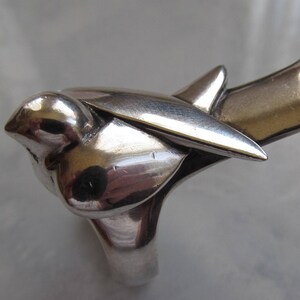 May include: A silver ring shaped like a bird in flight. The bird's wings are spread and its head is tilted down. The ring is made of polished metal and has a smooth, shiny finish.