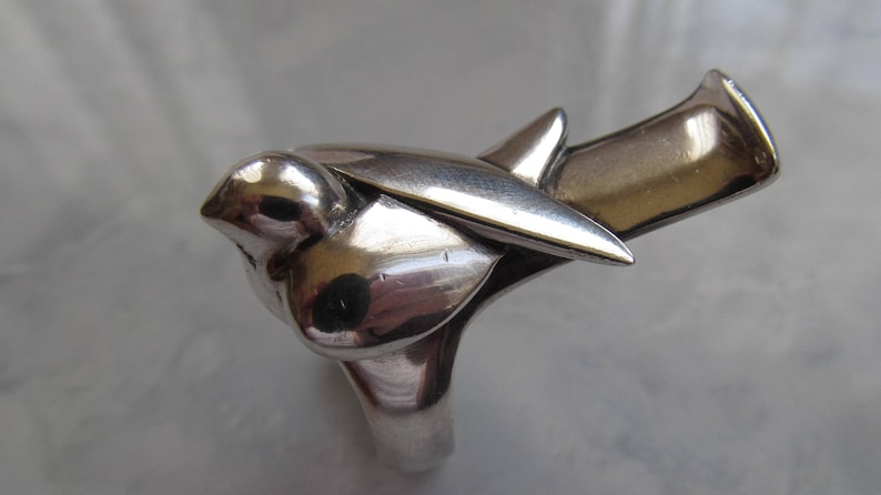 May include: A silver ring shaped like a bird in flight. The bird's wings are spread and its head is tilted down. The ring is made of polished metal and has a smooth, shiny finish.
