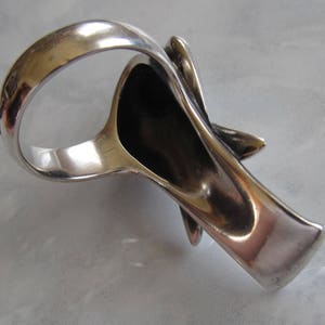 May include: A silver ring with a unique, abstract design. The ring is open on one side and has a textured, organic shape.
