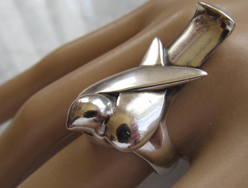 May include: A silver ring featuring a stylized bird design. The bird is depicted in flight with its wings spread wide. The ring is made of solid silver and has a smooth, polished finish.