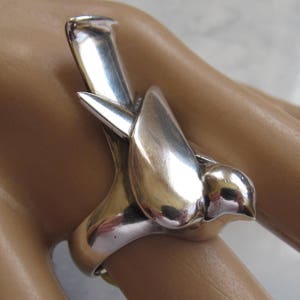 May include: A silver ring featuring a stylized bird design. The bird is depicted in flight with its wings outstretched. The ring is made of polished silver and has a smooth, rounded surface.