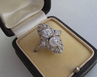 May include: A vintage diamond ring with two large round diamonds set in a platinum setting. The ring has a delicate, intricate design with smaller diamonds surrounding the larger stones.