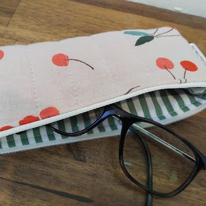 May include: A light pink quilted glasses case with a cherry pattern. The case has a white zipper and a green and white striped interior. Black eyeglasses are partially visible inside the case.
