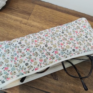 May include: A floral eyeglasses case with a white background and a zipper. The case is open, revealing a pair of black-framed glasses. The floral pattern includes pink, blue, and gray flowers.