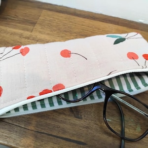 Op de afbeelding: Een rechthoekig gewatteerd stoffen etui met een lichtroze achtergrond, een print van rode kersen en groene blaadjes. Het etui heeft een witte ritssluiting en een groen-wit gestreepte voering, met een donkerbruine bril die er bovenop rust.