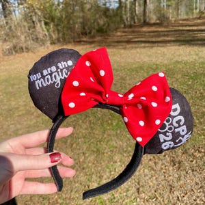 May include: Black mouse ear headband with a red bow featuring white polka dots. One ear has the text "You are the magic" and the other ear has the text "DCP 26".