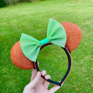 May include: Orange sequined mouse ears with a green bow and black headband. The bow is made of a sheer, glittery fabric. The ears are round and have a textured, glittery appearance. The headband is black with a black trim.