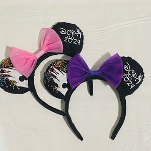 May include: A pair of black mouse ears with a pink and purple bow. The ears are embroidered with fireworks and the text "Disney 2024".