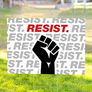 May include: A white yard sign with the word "RESIST" in red and gray, and a black fist graphic. The sign is displayed on a green lawn with a metal stake.