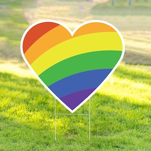 May include: A rainbow heart-shaped yard sign with a white background. The heart is made of red, orange, yellow, green, blue, indigo, and violet stripes.
