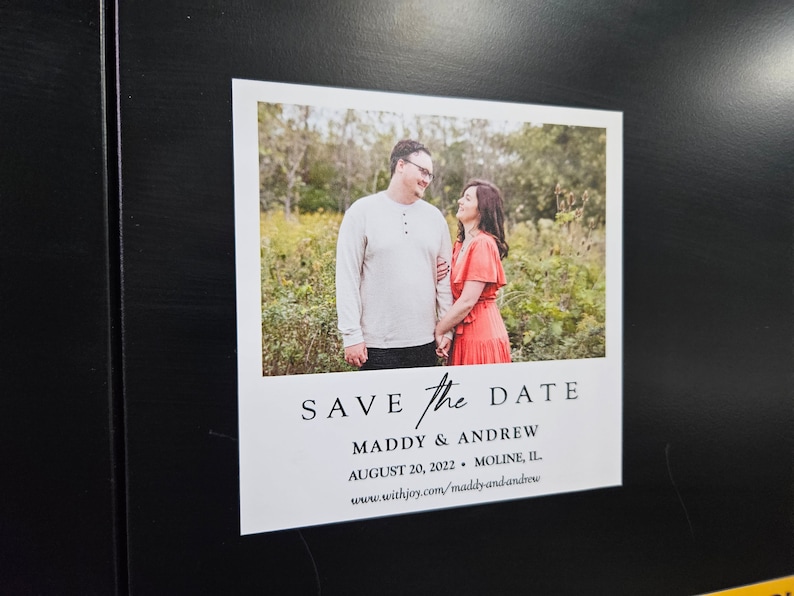 May include: A printed "Save the Date" card featuring a couple holding hands in a field. The card has the text "SAVE the DATE" at the top, followed by names, date, and location. The card is rectangular with a white border.