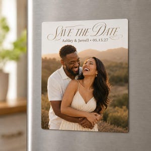 May include: A rectangular magnet featuring a photograph of two people embracing outdoors at sunset. The text "SAVE THE DATE" is printed in a dark script font at the top, followed by the names "Ashley & Jerrell" and the date "05.15.27".
