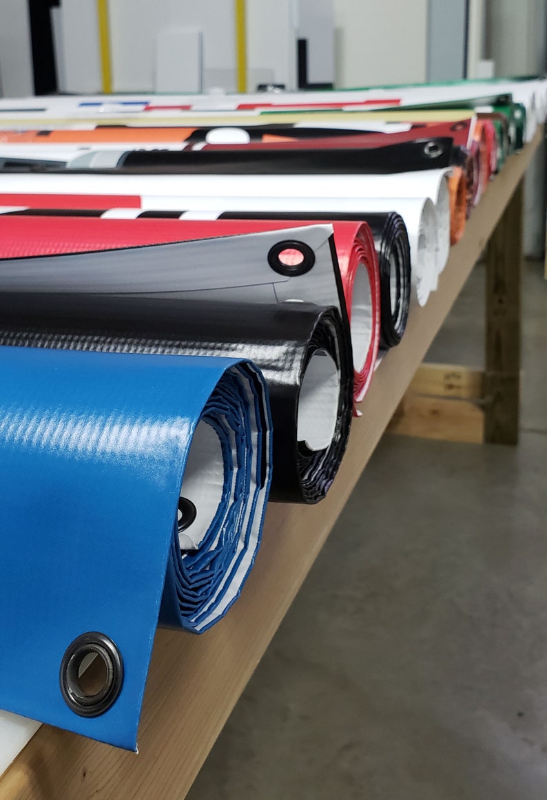 May include: A close-up of several rolled-up banners in various colors, including blue, black, red, and white. The banners are stacked on a wooden surface and have grommets along the edges.
