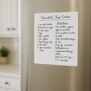 May include: A white rectangular magnet with handwritten chocolate chip cookie recipe details, including ingredients and directions, is affixed to a stainless steel refrigerator door. The text is in black ink.