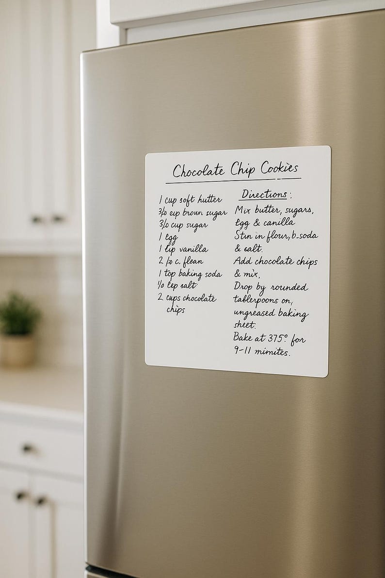 May include: A white rectangular magnet with handwritten chocolate chip cookie recipe details, including ingredients and directions, is affixed to a stainless steel refrigerator door. The text is in black ink.