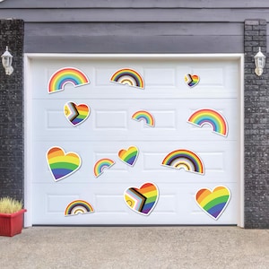 May include: A white garage door decorated with rainbow and heart-shaped decals. The decals feature a variety of rainbow colors and designs, including a pride flag and a progress pride flag.