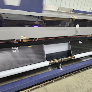 May include: A large format printer with two rolls of fabric being fed through the machine. The fabric is black with white text that reads "SE" and "10".