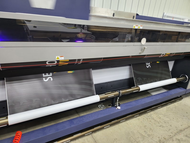 May include: A large format printer with two rolls of fabric being fed through the machine. The fabric is black with white text that reads "SE" and "10".