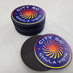 May include: A stack of circular magnets and one individual magnet featuring a dark blue background with a central orange and yellow spiral graphic. The text CITY SC and CHULA VISTA is printed in white capital letters around the spiral design.
