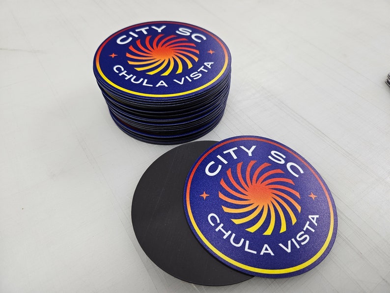 May include: A stack of circular magnets and one individual magnet featuring a dark blue background with a central orange and yellow spiral graphic. The text CITY SC and CHULA VISTA is printed in white capital letters around the spiral design.