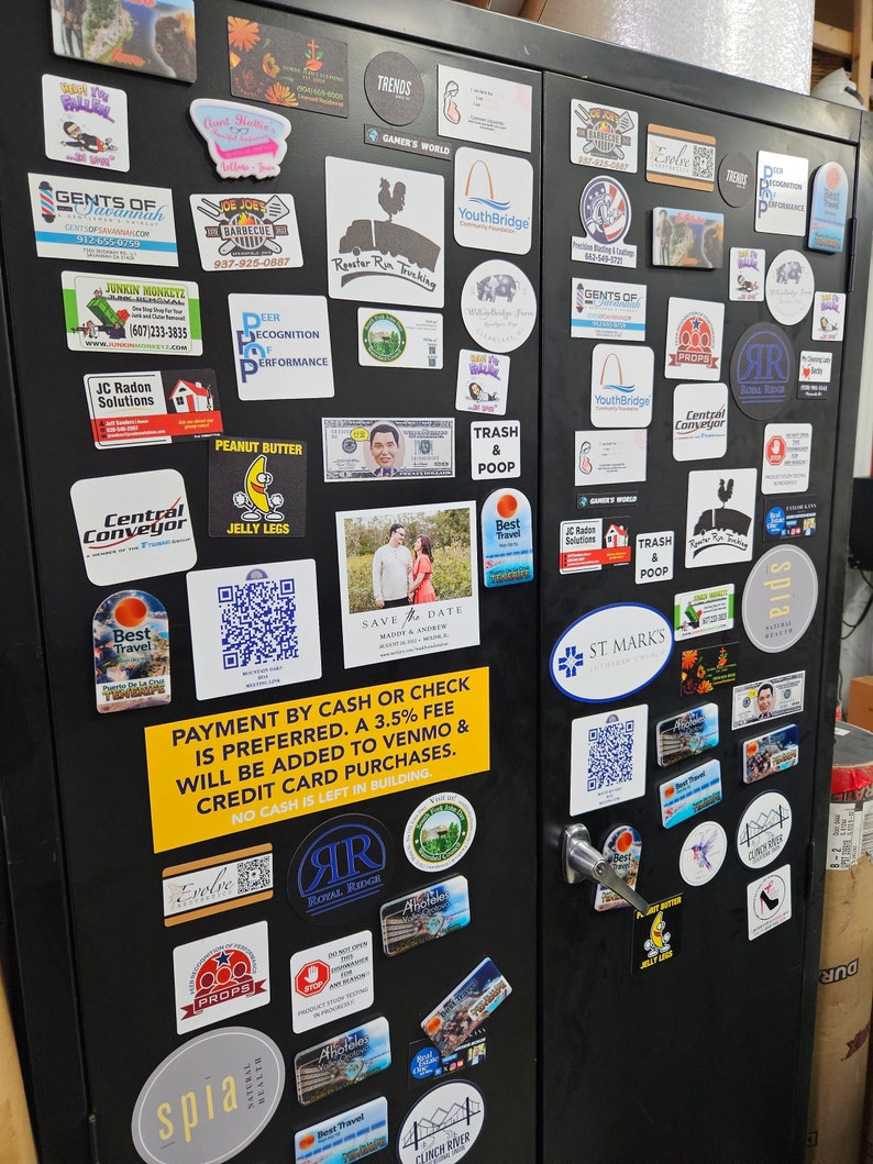 May include: A black metal cabinet covered in various stickers and business cards. The stickers feature logos, text, and graphics for different businesses and organizations. Some stickers have QR codes. A yellow sign states payment preferences.