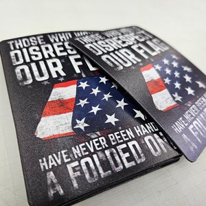 May include: Two rectangular metal signs featuring a graphic of the American flag and the text: THOSE WHO DISRESPECT OUR FLAG HAVE NEVER BEEN HANDED A FOLDED ONE. The design uses a distressed, weathered aesthetic with bold white lettering on a black background.