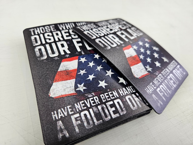 May include: Two rectangular metal signs featuring a graphic of the American flag and the text: THOSE WHO DISRESPECT OUR FLAG HAVE NEVER BEEN HANDED A FOLDED ONE. The design uses a distressed, weathered aesthetic with bold white lettering on a black background.