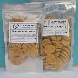 May include: Two clear plastic bags filled with pumpkin dog treats. The bags have white labels with the text "Pumpkin Dog Treats" and "La Barkeria". The treats are a light brown color and appear to be round and rectangular.