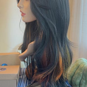 May include: A black wig with long, straight hair and subtle orange and yellow highlights. The wig is styled with a side part and is worn on a mannequin head.