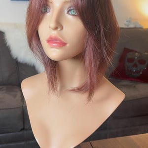 May include: A mannequin head with a short, layered wig in a reddish-brown hue. The mannequin has fair skin, green eyes, and glossy pink lips. It is mounted on a black and silver stand, suitable for displaying wigs or hairpieces.