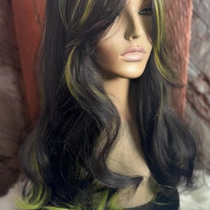 May include: A long, black wig with green highlights. The wig is styled in loose waves and has a side part. The wig is on a mannequin head.