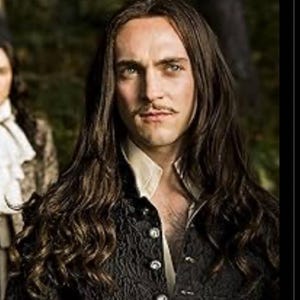 May include: A man with long, dark brown hair wearing a black jacket with silver buttons over a white shirt. He has a mustache and a serious expression.