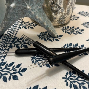 May include: Several black crochet hooks with silver tips are arranged on a white cloth with a blue leaf pattern. A light blue starfish and a silver mercury glass candle holder are in the background.