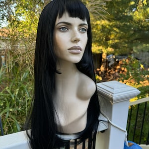 May include: A black wig with long straight hair and blunt bangs. The wig is styled on a mannequin head.